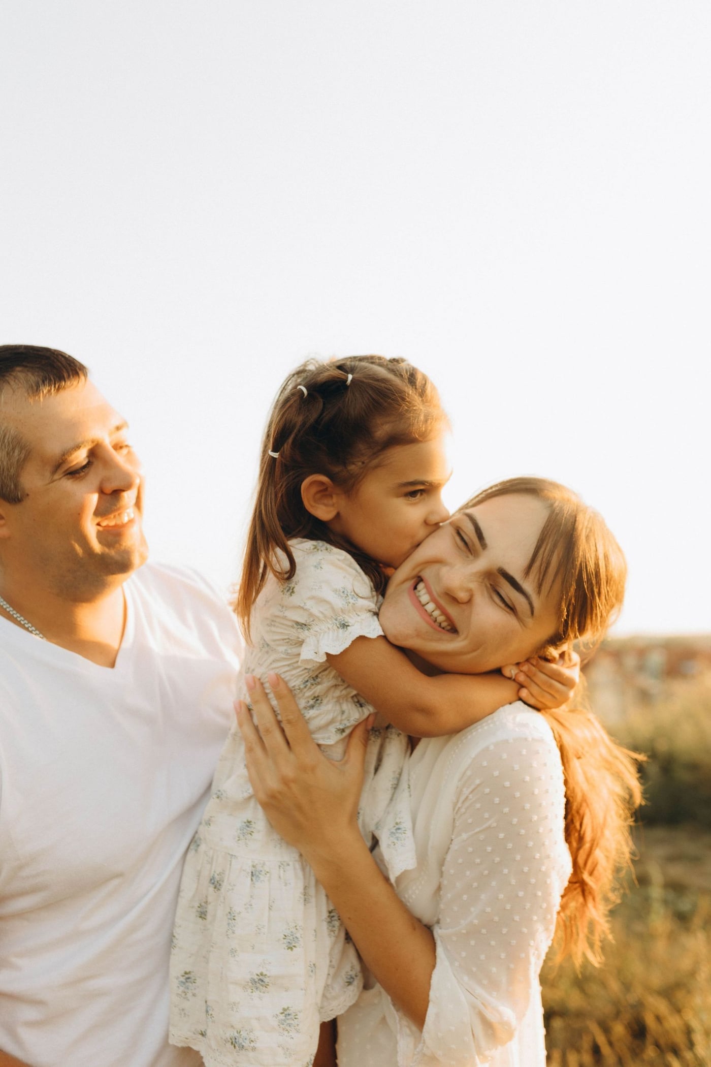 Família sorrindo ao ar livre sob luz dourada — pais e filha abraçados, transmitindo cuidado e proximidade.